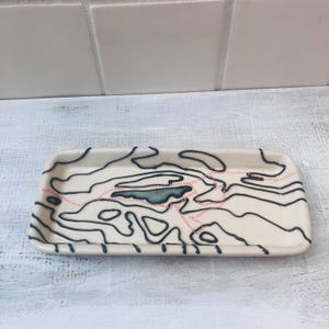 May include: A rectangular ceramic tray with a cream-colored base. The tray features a black, blue, and pink topographical design. The design includes black contour lines, a blue lake-like shape, and pink dotted lines.