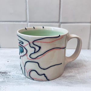 May include: A ceramic mug with a cream-colored exterior featuring dark blue hand-painted topographical lines and dotted coral-colored paths. The interior of the mug is a pale mint green, and it includes a curved handle on the right side.
