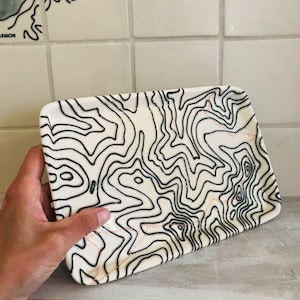 May include: A rectangular, off-white ceramic tray with a black topographical map design. The map features contour lines and a dotted red line. The tray is held by a hand, showcasing its size and shape. The design is a modern take on a classic map.