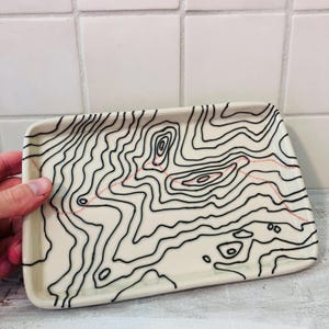 May include: A rectangular ceramic tray with a cream-colored base featuring a black topographic map pattern of contour lines and a single dotted red line running across the surface. The tray has rounded corners and a smooth, glossy finish.