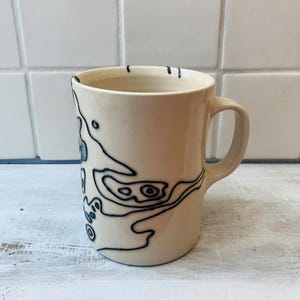 May include: Cream-colored ceramic mug with a black abstract design. The mug has a rounded body, a handle, and a glossy finish. The black design features organic shapes and lines, creating a unique, artistic look. The mug is on a white surface.