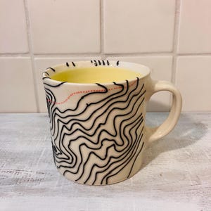 May include: A cream-colored ceramic mug featuring a raised black topographic map pattern of winding lines across the exterior surface. A single dotted orange line curves across the upper portion of the mug, which has a pale yellow interior.