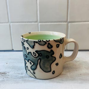 May include: Cream-colored ceramic mug with a blue and black abstract design. The mug has a rounded handle and is filled with a light green liquid. The mug is sitting on a white wooden surface.
