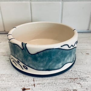 May include: A ceramic bowl with a cream-colored interior and a teal and white exterior design. The bowl has a dark blue base and is decorated with black wavy lines and abstract shapes. The bowl is likely handmade.