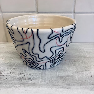 May include: A white ceramic bowl with a topographic map design. The map features navy blue contour lines and dotted pink lines. The bowl has a slightly flared shape and a smooth, glossy finish. The interior is a creamy off-white color.
