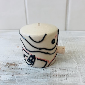 May include: A ceramic bottle stopper with a cork base. The off-white stopper is decorated with black, wavy lines and small red dots. The design is abstract and artistic, suitable for wine bottles.