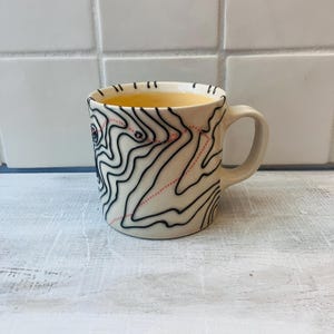 May include: Cream-colored ceramic mug with a black and red topographic map design. The mug has a curved handle and is filled with a light yellow liquid. The map design features black contour lines and a dotted red line.