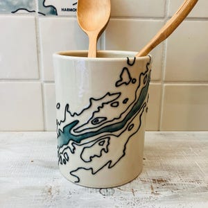 May include: A cream-colored ceramic utensil holder with a teal and black abstract design. Two wooden spoons are inside the holder. The holder is cylindrical and sits on a white surface.