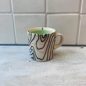 May include: A ceramic mug with a cream-colored base, featuring a black topographical design and a dotted pink line. The mug is filled with a light green liquid. The mug has a handle and is set against a white tile background.