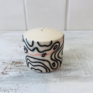 May include: A cream-colored ceramic salt shaker with a cylindrical shape. The shaker is decorated with black, wavy lines and a dotted pink line. The top has two small holes for dispensing salt.