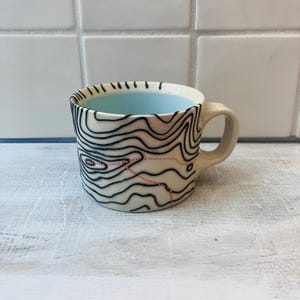 May include: Cream-colored ceramic mug with a black topographical map design. The interior is light blue. A red dotted line and a small red area are part of the map design. The mug has a curved handle.