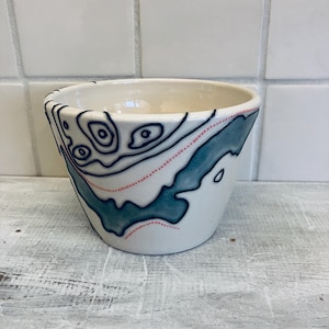 May include: A white ceramic bowl with a blue and teal topographical design. The bowl features a map-like pattern with dark blue lines, teal shapes, and dotted pink lines. The bowl is on a white wooden surface.