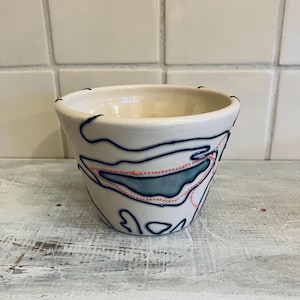 May include: A small, off-white ceramic bowl with a rounded shape. The bowl is decorated with blue and red abstract designs. The rim of the bowl has small black accents. The bowl sits on a distressed, white wooden surface.