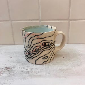 May include: Cream-colored ceramic mug with a black topographical map design and a light blue interior. The map features black contour lines and pink dotted lines. The mug has a curved handle and sits on a white surface.