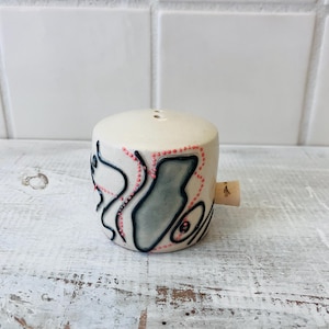 May include: A ceramic salt shaker with a cork stopper. The off-white shaker features a design of black and gray shapes outlined with pink dots and lines. Two small holes are on the top of the shaker.