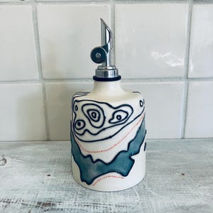May include: A ceramic oil bottle with a stainless steel spout. The off-white bottle features a hand-painted design with black, teal, and red accents. The design resembles a topographic map. A stylish kitchen accessory.