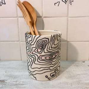 May include: A cylindrical ceramic utensil holder with a cream-colored base featuring black topographic map contour lines and a thin red dashed line pattern. Two light brown wooden utensils are placed inside the container.