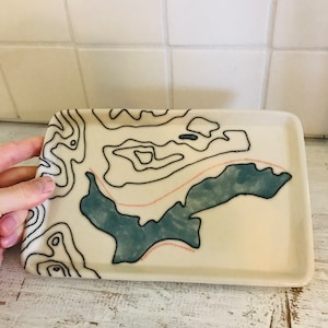 May include: A white ceramic tray with a topographic map design. The map features black lines and a blue-green area with a pink dotted line.