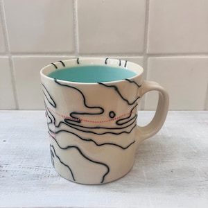 May include: Cream-colored ceramic mug with a black topographical design and a pink dotted line. The interior of the mug is a bright turquoise color. The mug has a curved handle and is set against a white background.