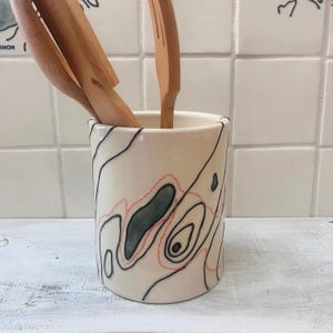 May include: A cylindrical ceramic utensil holder with a cream-colored base, decorated with black contour lines and red dotted lines. Wooden kitchen utensils are inside the holder. The design resembles a topographic map.