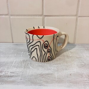 May include: A ceramic mug with a cream-colored exterior featuring black topographic contour lines and a dotted red trail pattern. The interior of the mug is a solid, vibrant red. It has a curved handle on the right side and a smooth finish.