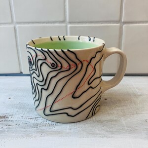 May include: Cream-colored ceramic mug with a black topographical map design and a light green interior. The map features black contour lines and red dotted lines. The mug has a curved handle.