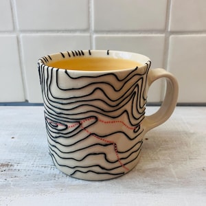 May include: Cream-colored ceramic mug with a black topographical map design. The mug has a curved handle and is filled with a yellow liquid. The map design includes a dotted red line. The mug is on a white surface.