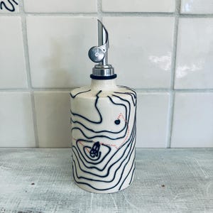 May include: A ceramic oil bottle with a stainless steel spout. The bottle is off-white with a black topographical map design and a red dotted line. The spout has a small, round, silver-colored lever.