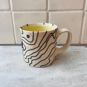May include: Cream-colored ceramic mug with a black topographical map design. The mug has a curved handle and is filled with a yellow liquid. The map design includes a dotted red line and a black dot.