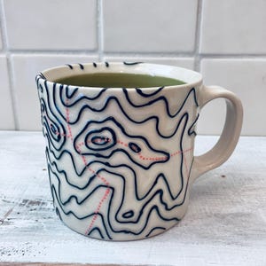 May include: A ceramic mug with a cream-colored exterior featuring raised dark blue topographic contour lines and dotted coral-colored paths. The interior of the mug is a solid light green, and it includes a curved handle on the right side.