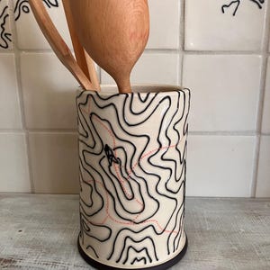 May include: A ceramic utensil holder with a black and white topographic map design and a red dotted line. Two wooden spoons are inside the holder. The base of the holder is black, and the background features white tiles with the word "PEEKSKILL".