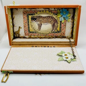 May include: Open wooden box with an orange frame, featuring a leopard illustration and floral accents. The interior has a patterned design, with a giraffe and the word "STERLING" visible. A chain and flower embellishment are attached.