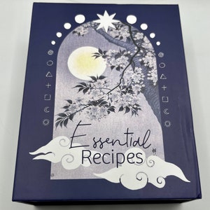 Könnte beinhalten: Eine marineblaue Box mit einer weißen und grauen Illustration eines Mondes, Sternen und eines Baumes mit weißen Blüten. Der Text "Essential Recipes" ist in weißer Kursivschrift geschrieben.
