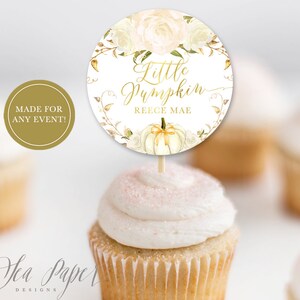 Könnte beinhalten: Ein weißer Cupcake mit goldenen Glitzer oben und einem runden Topper, auf dem "Little Pumpkin Reece Mae" mit einem weißen Kürbis und goldenen Blättern steht.