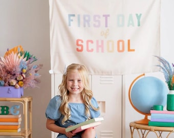 Könnte beinhalten: Ein junges Mädchen in einem blauen Kleid sitzt auf einem Hocker und hält einen Stapel Bücher. Sie lächelt in die Kamera. Hinter ihr befindet sich ein weißer Schrank mit einem Banner, auf dem "FIRST DAY of SCHOOL" in Regenbogenfarben steht. Auf einem Tisch stehen auch Bücher und ein Globus.