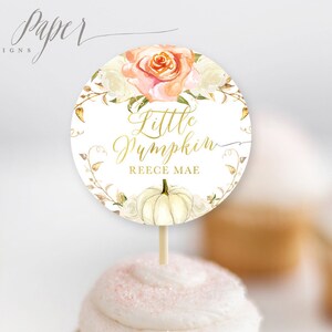 May include: A round, white and gold cupcake topper with a watercolor design of a pumpkin, flowers, and leaves. The text on the topper reads "Little Pumpkin Reece Mae".
