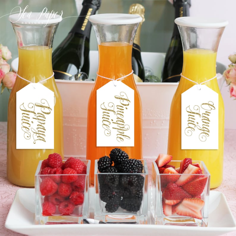 May include: Three glass carafes filled with orange, yellow, and light orange juice. Each carafe has a white tag with gold lettering indicating the type of juice: "Orange Juice", "Pineapple Juice", and "Papaya Juice".  There are three small glass bowls filled with fresh fruit: strawberries, blackberries, and raspberries.