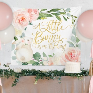 May include: A white banner with a watercolor floral design and a gold inscription that reads "A little bunny is on the way!"  The banner is decorated with pink and white flowers, green leaves, and a cute bunny illustration.