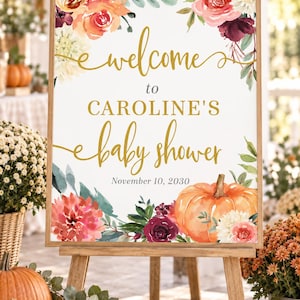 May include: A rectangular sign in a wooden frame featuring the text "welcome to CAROLINE'S baby shower November 10, 2030" in gold and black lettering, decorated with watercolor illustrations of orange pumpkins and various red and white flowers.
