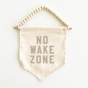 May include: A beige canvas banner with the text "NO WAKE ZONE" in brown lettering. The banner is hanging from a wooden dowel with a string of wooden beads.