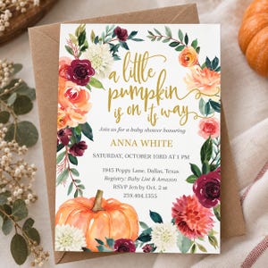 May include: A rectangular baby shower invitation card featuring a watercolor floral wreath in shades of burgundy, orange, and white, with a large orange pumpkin at the bottom. The gold text reads: A little pumpkin is on its way, Anna White.