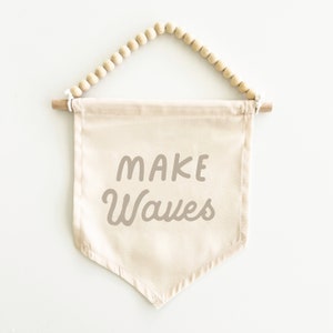 May include: A beige canvas banner with wooden dowels and a beaded string hanger. The banner has the text "MAKE WAVES" printed in a brown cursive font.
