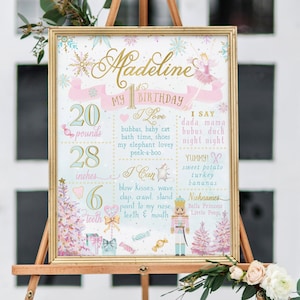 May include: A pink and blue first birthday sign for a girl named Madeline. The sign features a ballerina, a nutcracker, and a Christmas tree. It includes details like her weight (20 pounds), height (28 inches), and number of teeth (6). The sign also lists things she loves, can do, and says.