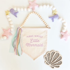 May include: A beige canvas banner with the text "MAKE WAVES LITTLE MERMAID" in pink lettering. The banner is decorated with pastel pink, blue, and green yarn tassels. The banner is hanging from a wooden beaded garland with white felt balls and pink and yellow felt stars.