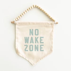 May include: A beige canvas banner with a wooden dowel and beaded string hanging system. The banner has the text "NO WAKE ZONE" printed in light blue.