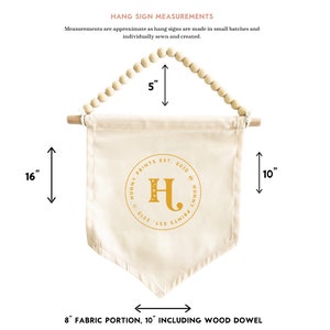 K&ouml;nnte beinhalten: Cremefarbenes Stoff-H&auml;ngeschild mit Holzstab und Perlenbesatz. Das Schild ist 40,6 cm lang und 20,3 cm breit, mit dem Text "HUNNY PRINTS EST. 2013" und einem goldenen Buchstaben "H".