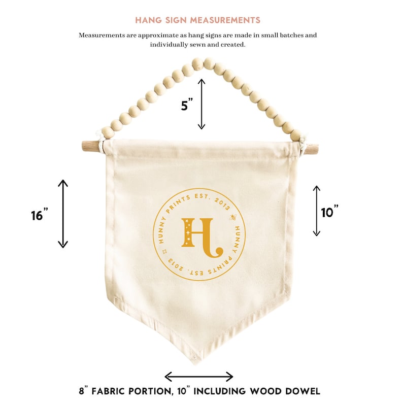 K&ouml;nnte beinhalten: Cremefarbenes Stoff-H&auml;ngeschild mit Holzstab und Perlenbesatz. Das Schild ist 40,6 cm lang und 20,3 cm breit, mit dem Text "HUNNY PRINTS EST. 2013" und einem goldenen Buchstaben "H".