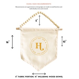 May include: Cream-colored fabric hang sign with a wooden dowel and beaded top. The sign is 16 inches long and 8 inches wide, with the text "HUNNY PRINTS EST. 2013" and a gold letter "H".