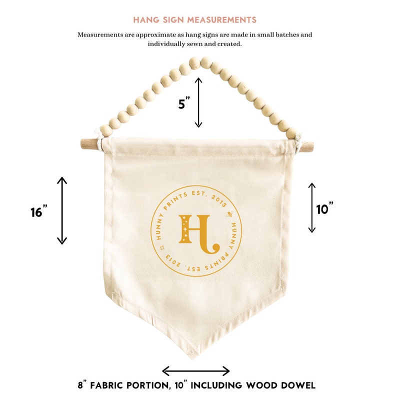 May include: Cream-colored fabric hang sign with a wooden dowel and beaded top. The sign is 16 inches long and 8 inches wide, with the text "HUNNY PRINTS EST. 2013" and a gold letter "H".