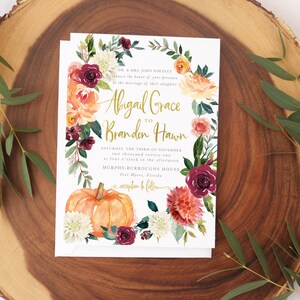 Op de afbeelding: Een huwelijksuitnodiging met een aquarelbloemenkrans, een pompoen en de tekst "Abigail Grace to Branden Hawn" in gouden letters. De uitnodiging ligt op een houten achtergrond.